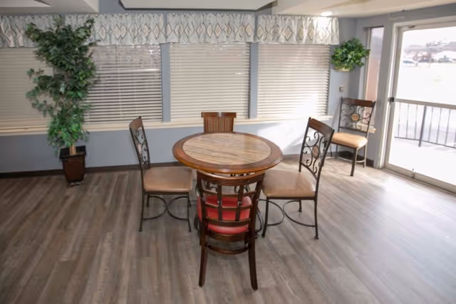 Small round dining table with four chairs in a bright common room with large windows, potted plants, and a glass door.