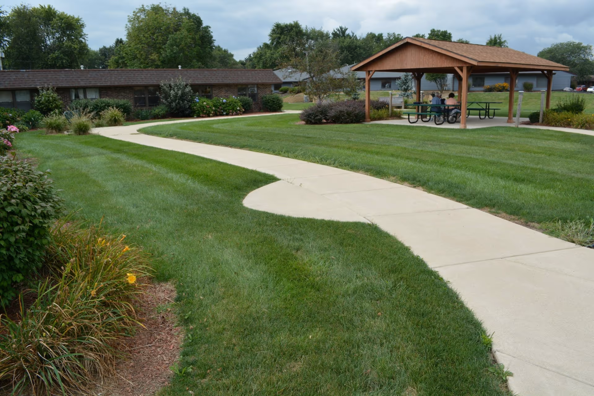 A well-maintained outdoor area at Valparaiso Care and Rehabilitation featuring a curved concrete walkway through green grass, landscaped bushes and flowers, a wooden pavilion with picnic tables, and two people sitting under the pavilion. Residential buildings and trees are visible in the background under a cloudy sky.