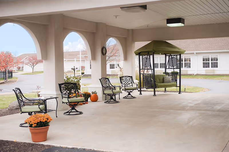 Covered outdoor seating area with metal chairs featuring green cushions and a green cushioned swing under a roof with arches. There are potted orange flowers and a pumpkin decoration on the concrete floor. In the background, there are white buildings and trees with autumn foliage.