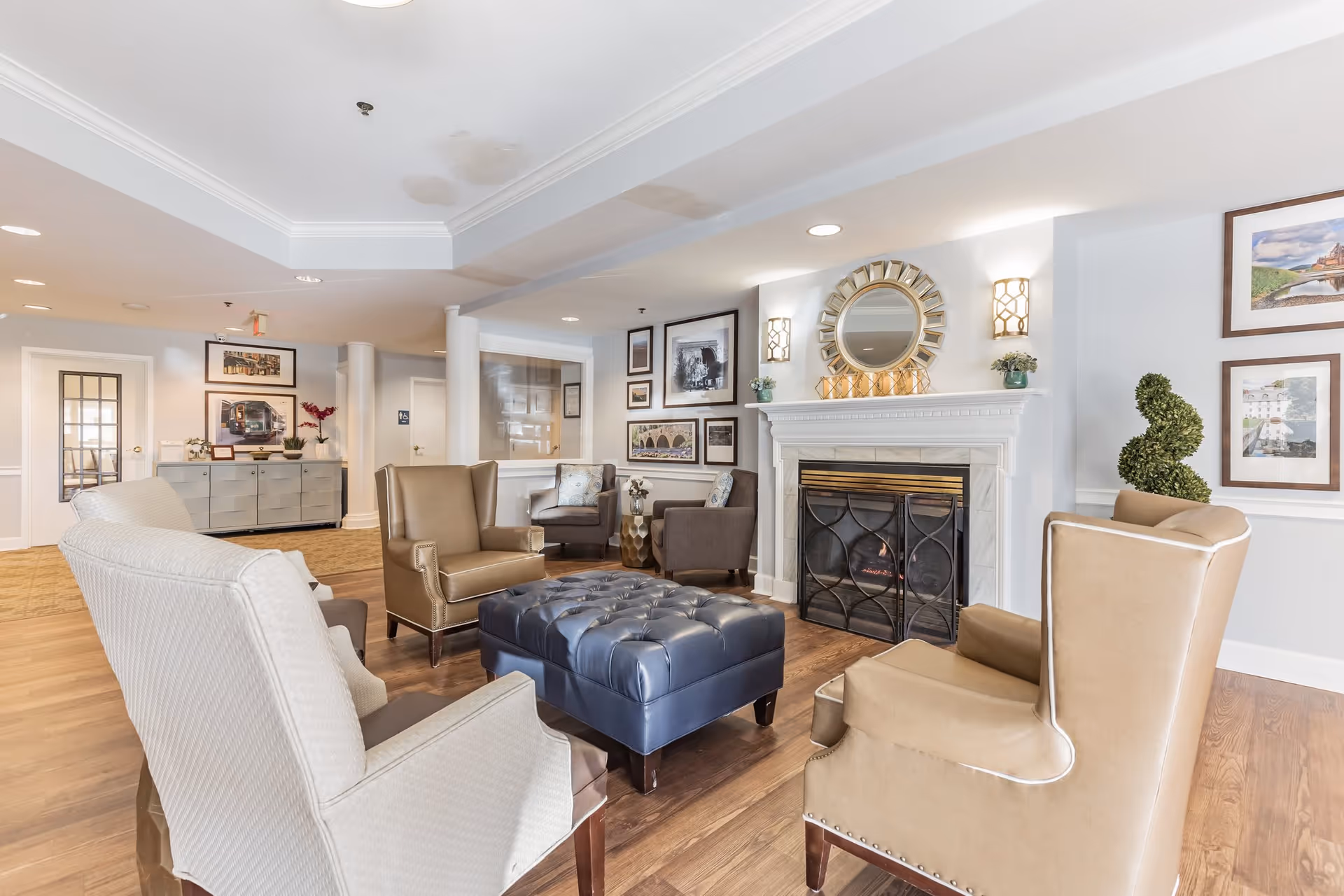 A cozy senior living facility lounge area with beige and brown armchairs arranged around a large blue tufted ottoman. There is a white fireplace with a decorative mirror and sconces above it, framed pictures on the walls, and wooden flooring. The space is well-lit with recessed lighting and has a welcoming atmosphere.