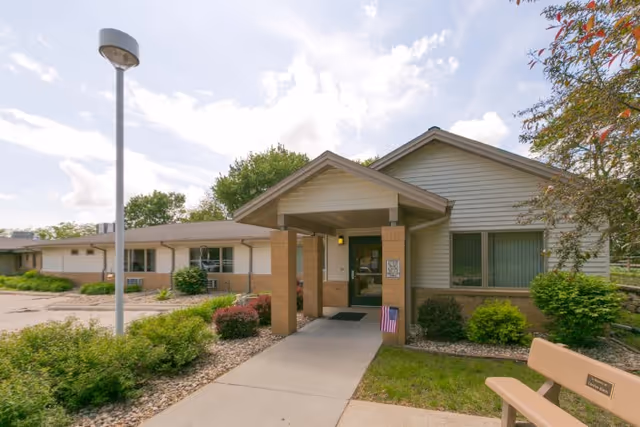 Exterior view of Careage Hills Rehabilitation And Healthcare facility showing the entrance with a covered porch supported by brick columns, a concrete walkway, surrounding greenery, a bench, and a streetlamp under a partly cloudy sky.
