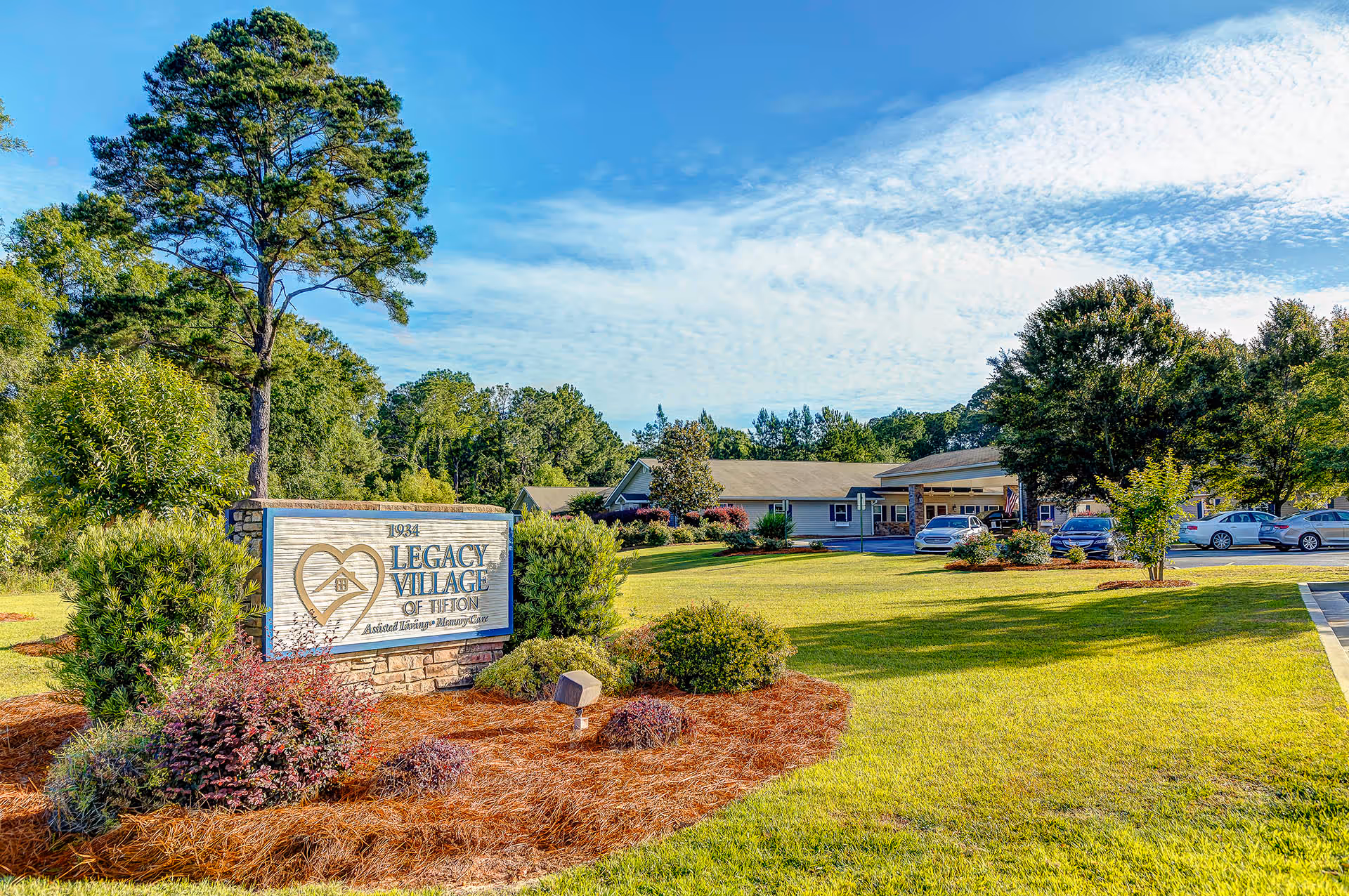 Exterior view of Legacy Village of Tifton, an assisted living and memory care facility, showing a landscaped lawn with bushes and trees, a stone sign with the facility name, and a single-story building with a parking area and several cars under a partly cloudy blue sky.