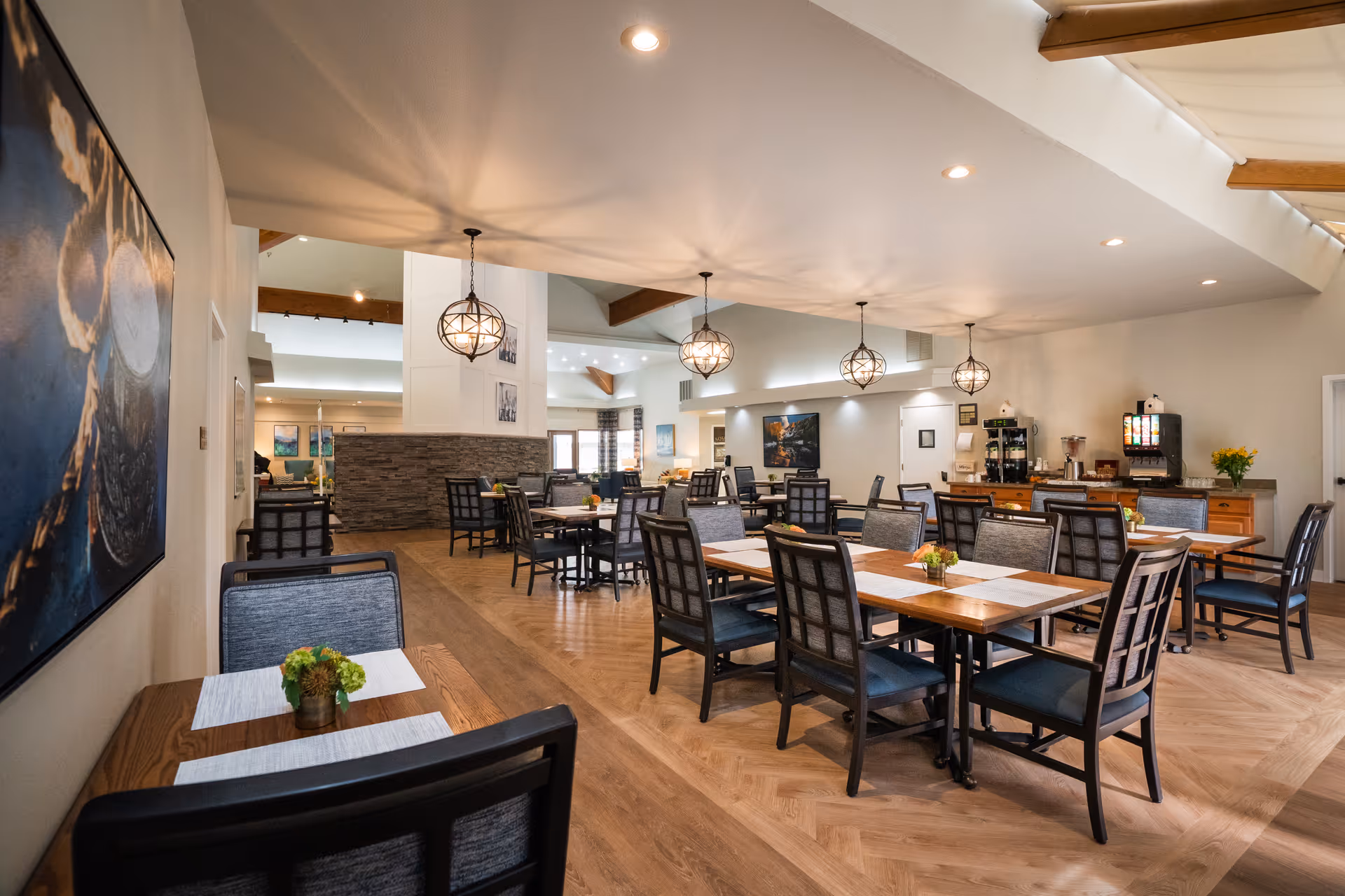 A spacious dining room with multiple wooden tables and cushioned chairs arranged neatly. The room features modern pendant lights hanging from the ceiling, a beverage station with coffee and soda machines along the back wall, and decorative plants on the tables. The flooring is wood with a herringbone pattern, and there is a large painting on the left wall.