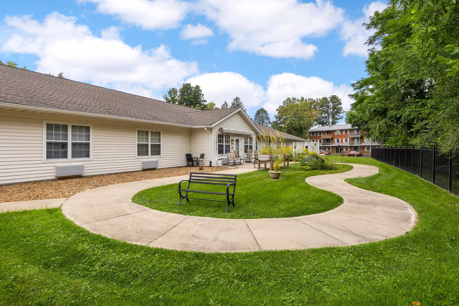 Curved concrete walkway and bench in a grassy courtyard outside a single-story senior living building with patio seating.