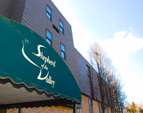 Exterior view of a multi-story building with a green awning that reads 'Shepherd of the Valley'. Leafless trees line the side of the building under a partly cloudy sky.