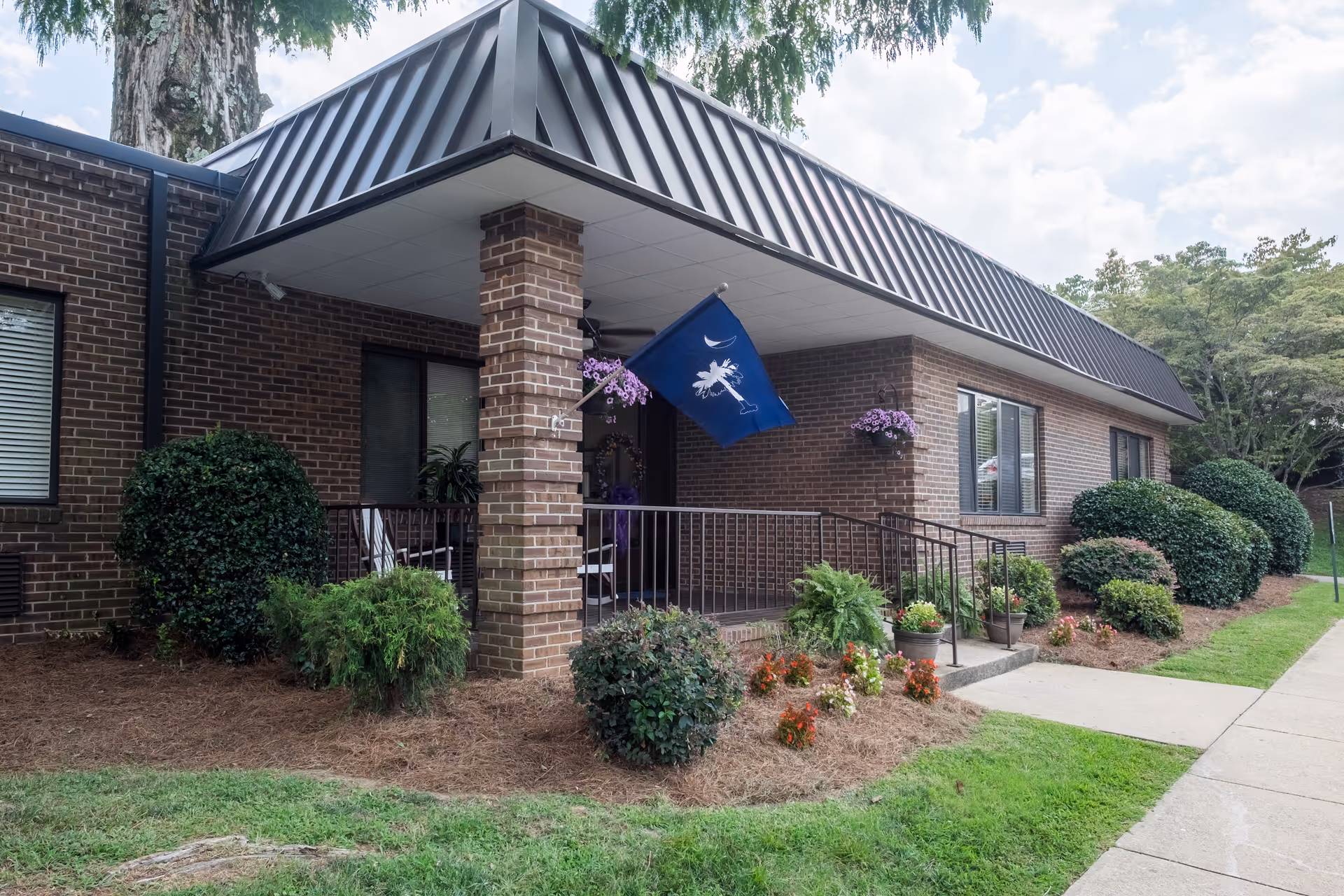 Brick single-story building entrance with a covered porch, landscaping, potted plants, and a South Carolina flag.
