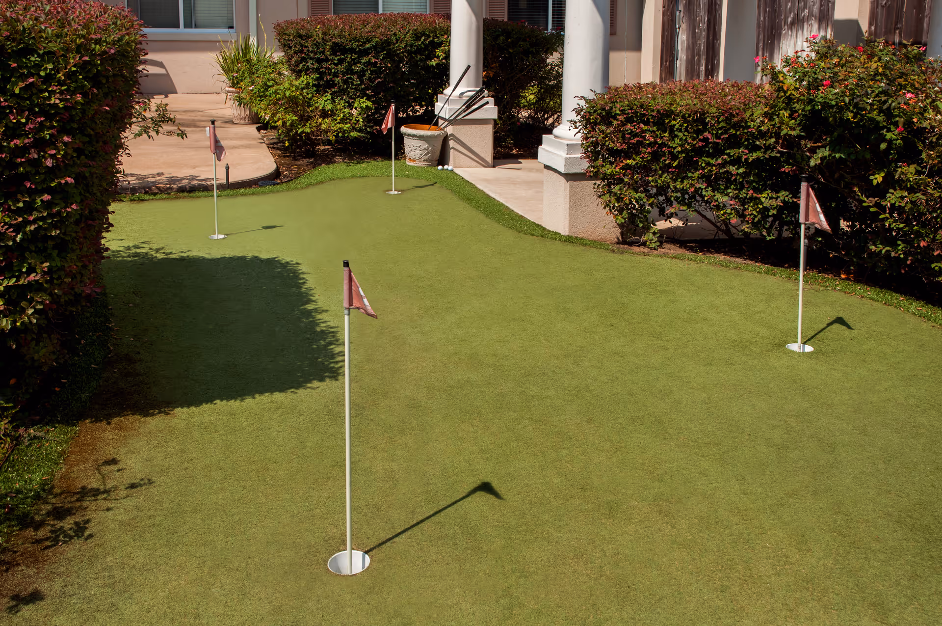 Small outdoor putting green with several flagsticks in front of a building entrance and surrounded by bushes.