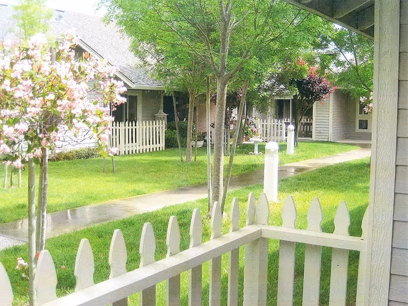 View of a green outdoor courtyard area in a retirement living community with a white picket fence in the foreground, trees, flowering plants, a paved walkway, and single-story buildings with porches in the background.