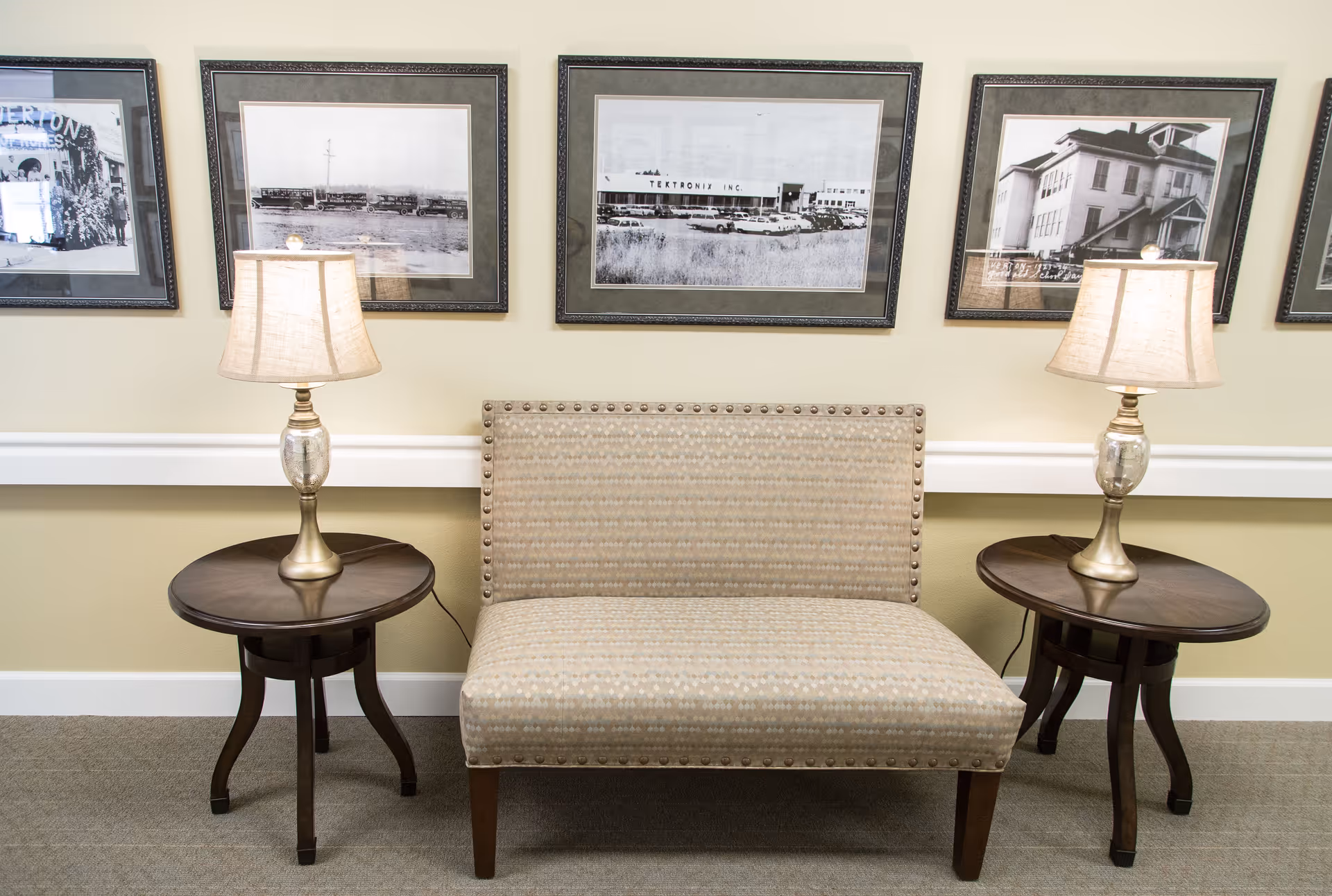 A small sitting area with a beige upholstered loveseat flanked by two round wooden side tables, each holding a table lamp with a beige shade. Behind the loveseat, there are four framed black and white photographs hanging on a light yellow wall.