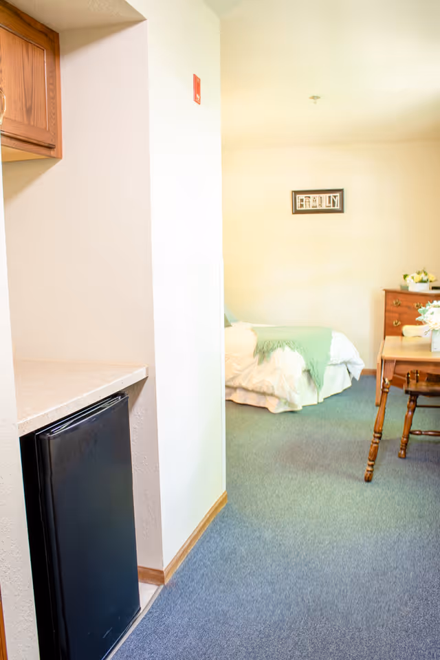 View of a senior living facility room showing a small kitchenette area with a black mini fridge and wooden cabinets on the left, leading into a bedroom area with a bed covered in white bedding and a green throw blanket. A wooden table with chairs and a dresser with flowers are visible in the background. A framed picture hangs on the wall above the bed.