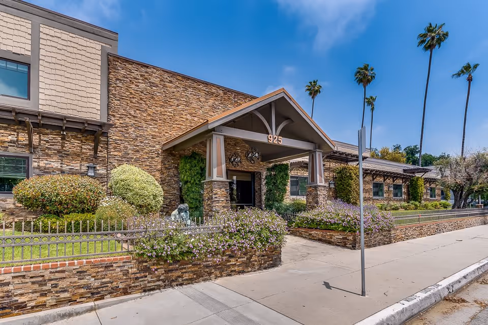 Exterior view of Astoria Park Senior Living building with stone facade, a covered entrance marked with the number 925, landscaped bushes and flowers, and tall palm trees under a clear blue sky.