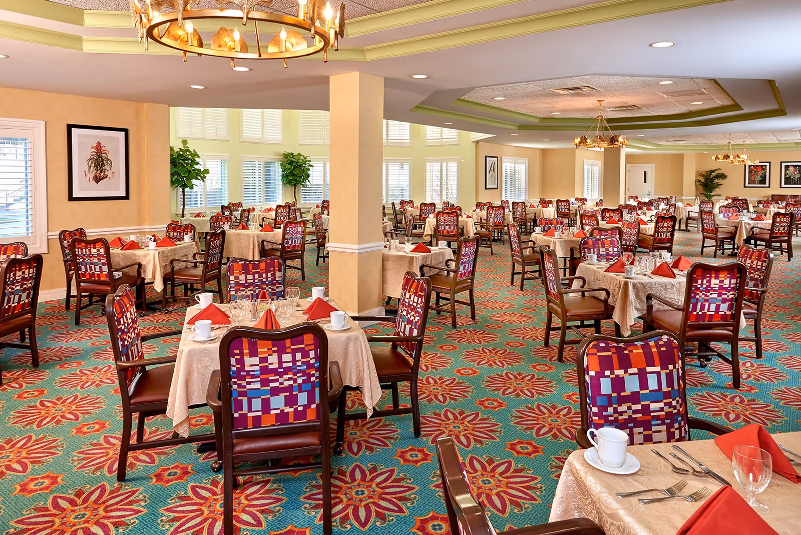 A spacious dining room with multiple tables covered in beige tablecloths, each set with white cups, glasses, silverware, and red folded napkins. The chairs have colorful patterned backs and brown leather seats. The room features large windows with white shutters, green plants, framed artwork on the walls, and decorative ceiling lights.