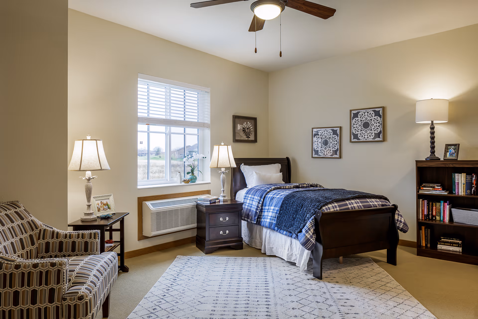 A cozy bedroom with a single dark wood bed dressed in blue and white plaid bedding and a dark blue throw blanket. There are two bedside tables with lamps on either side of the bed. A patterned armchair is positioned near a small table with a lamp and a framed photo. A window with white blinds lets in natural light, and a bookshelf filled with books and a lamp is against the wall. The room has beige walls, a ceiling fan with light, and a patterned area rug on the floor.