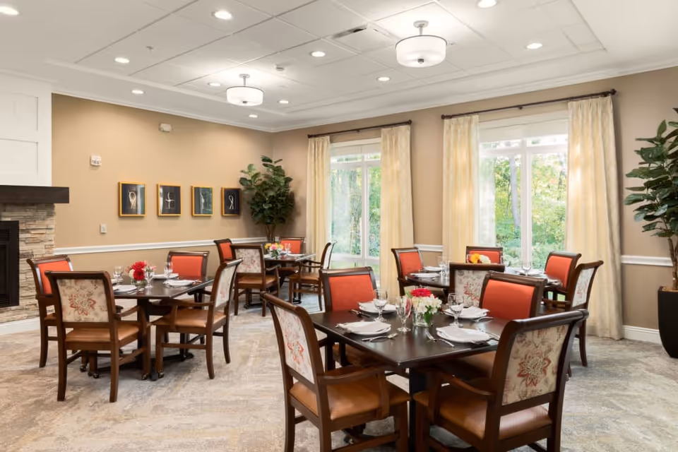 Dining room with several dark wood tables set for meals, upholstered chairs, large windows with curtains and potted plants.