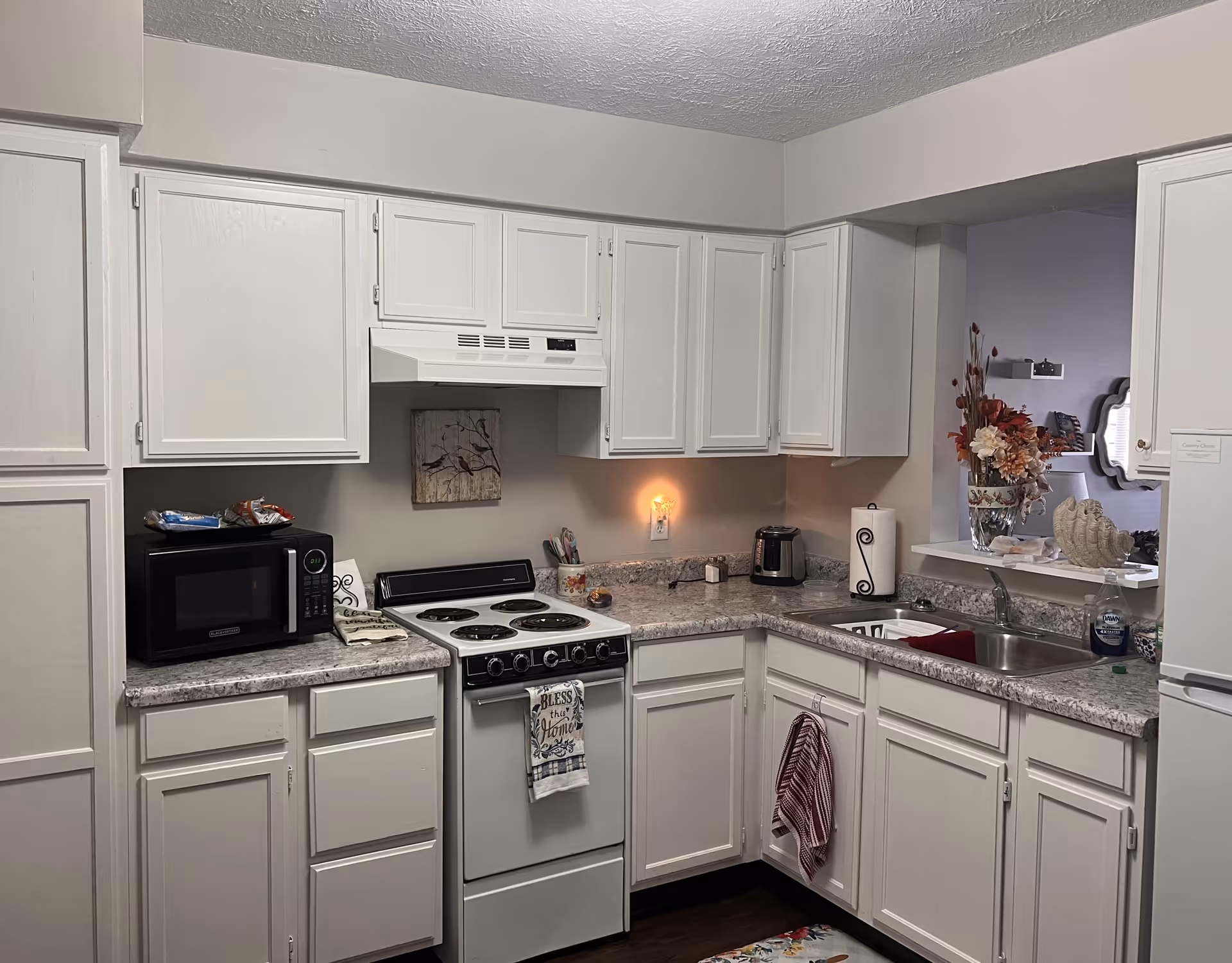 Compact kitchen with white cabinets, a stove and range hood, microwave, sink, and countertop appliances and decorations.