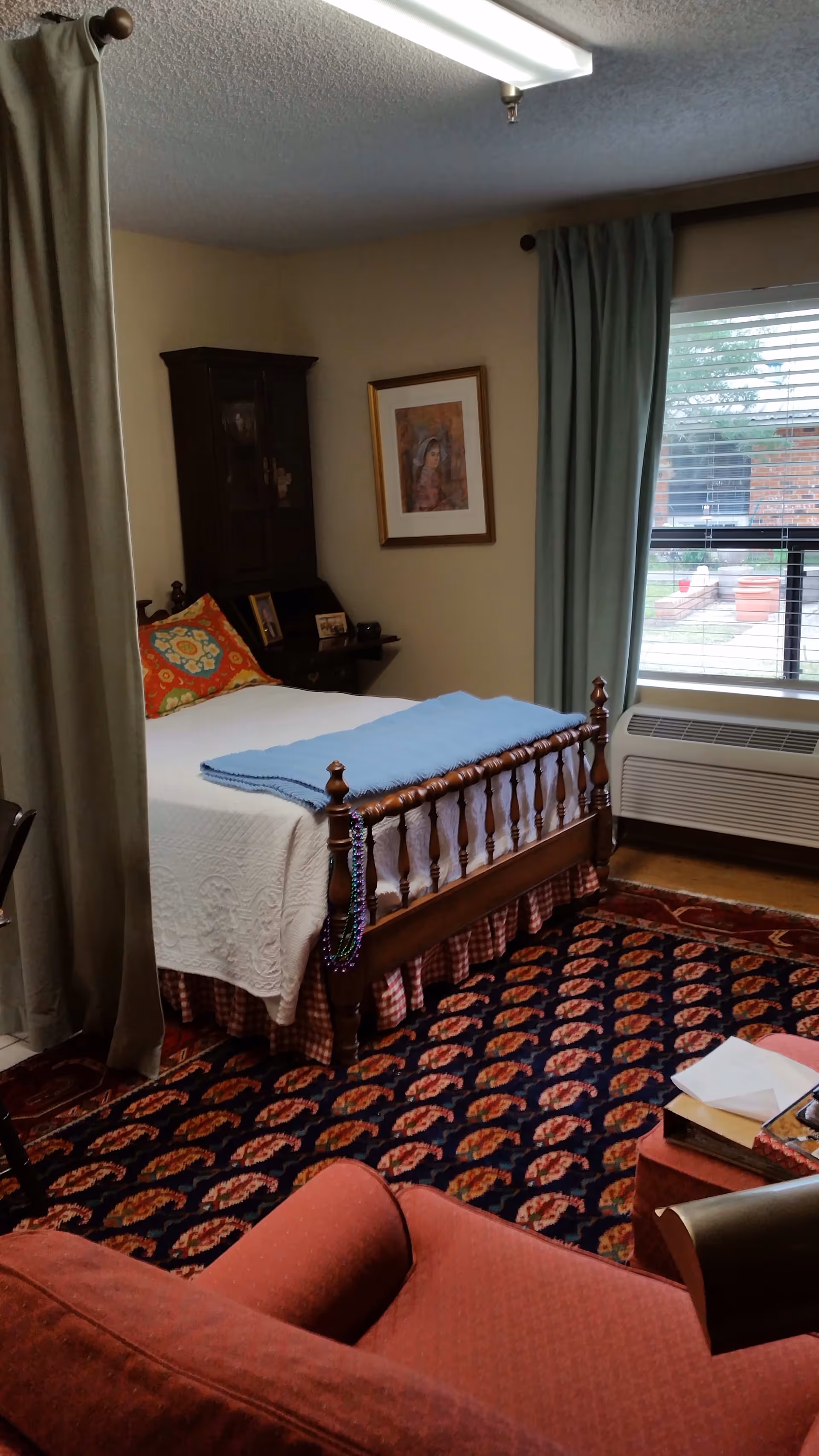 A cozy bedroom in a retirement community featuring a wooden bed with a white quilt and a blue blanket folded at the foot. There is a colorful pillow on the bed and a string of beads hanging from the bedpost. A dark wooden cabinet and framed artwork are on the wall. A window with green curtains lets in natural light, and a patterned carpet covers the floor. In the foreground, there is a red upholstered chair and a small table with books and papers.