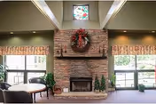 Spacious common room with a stone fireplace topped by a wreath, seating, plants, and large windows on either side.
