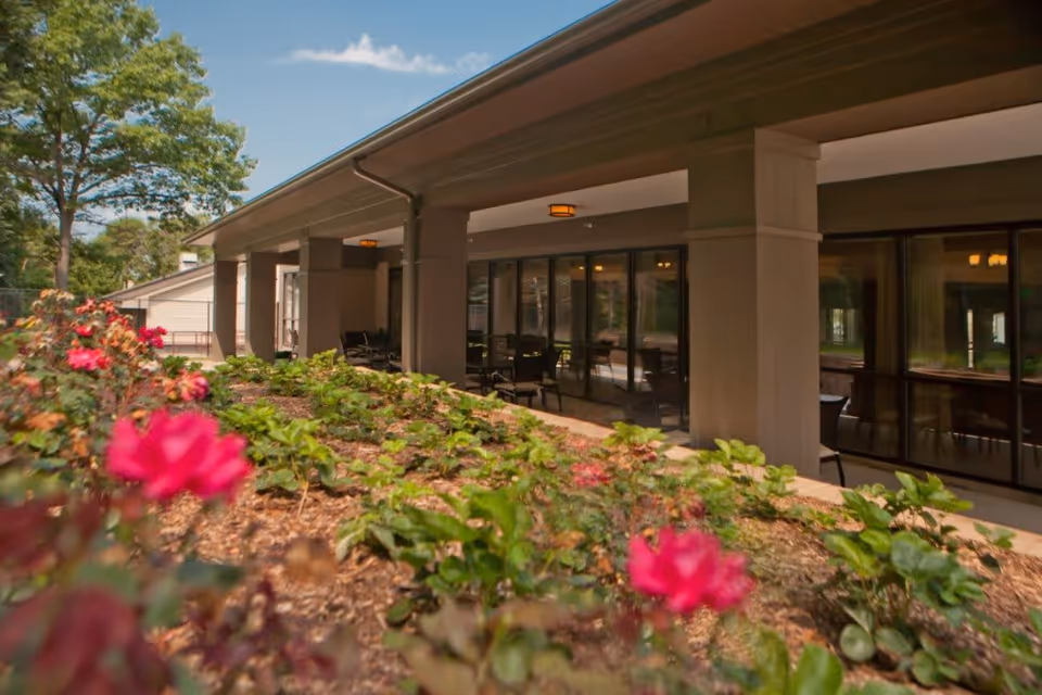 Outdoor view of a covered patio area with seating at The Cortland Northview, featuring a garden bed with blooming red flowers in the foreground and large windows along the building.
