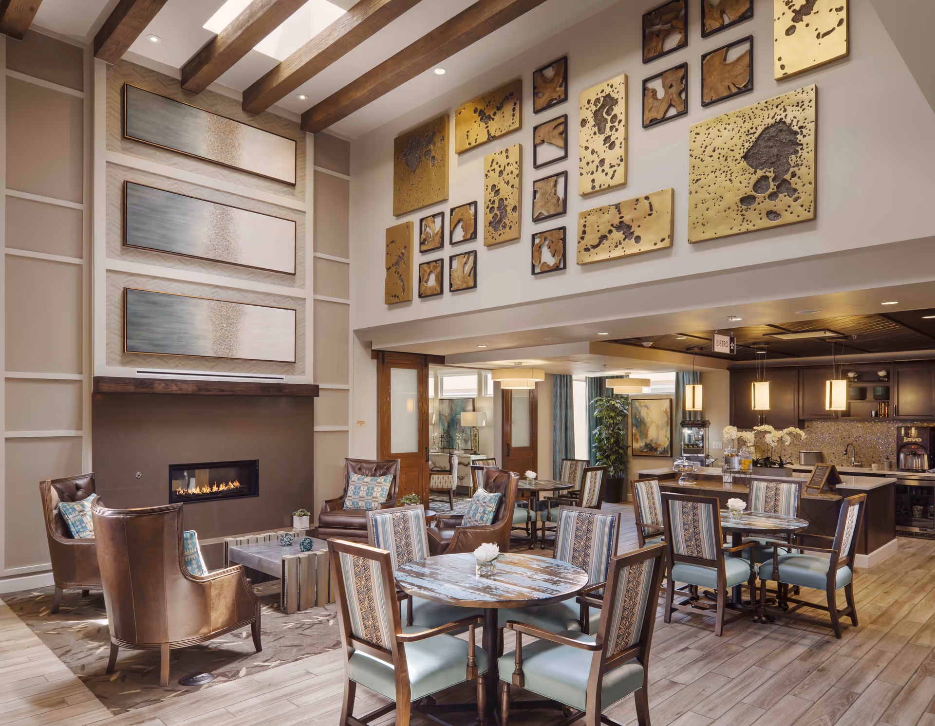 A spacious and well-lit common area in an assisted living facility featuring a modern fireplace with three abstract paintings above it, several round tables with chairs, and a kitchen area with pendant lights and a mosaic backsplash. The room has wooden ceiling beams and decorative wall art, creating a warm and inviting atmosphere.