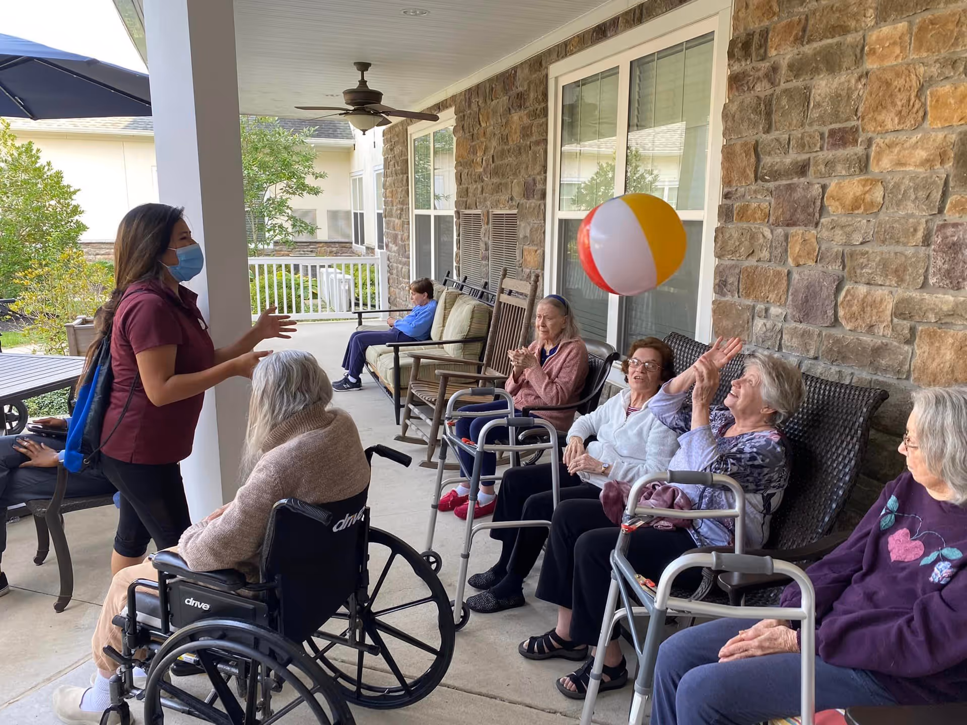 A group of elderly women sitting on chairs and wheelchairs on a covered outdoor patio, playing with a colorful beach ball. A caregiver wearing a mask is engaging with them. The patio has stone walls, ceiling fans, and overlooks greenery.