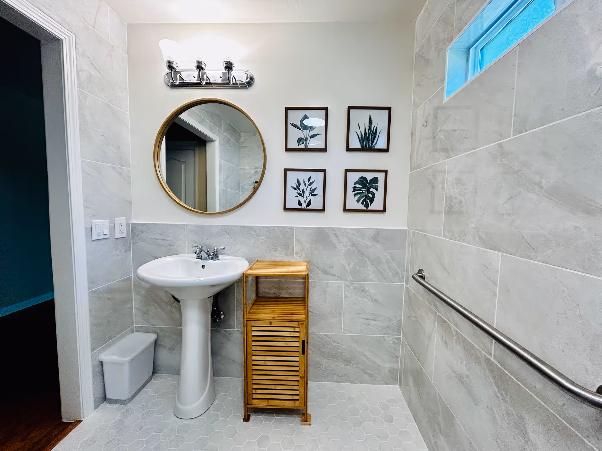 A clean bathroom with light gray tiled walls and hexagonal floor tiles. There is a white pedestal sink with a silver faucet, a round mirror with a wooden frame above it, and a wooden cabinet with slatted doors next to the sink. Four framed botanical prints hang on the wall above the cabinet. A metal grab bar is mounted on the right wall, and a small frosted window is near the ceiling. A white trash bin is placed on the floor near the doorway.