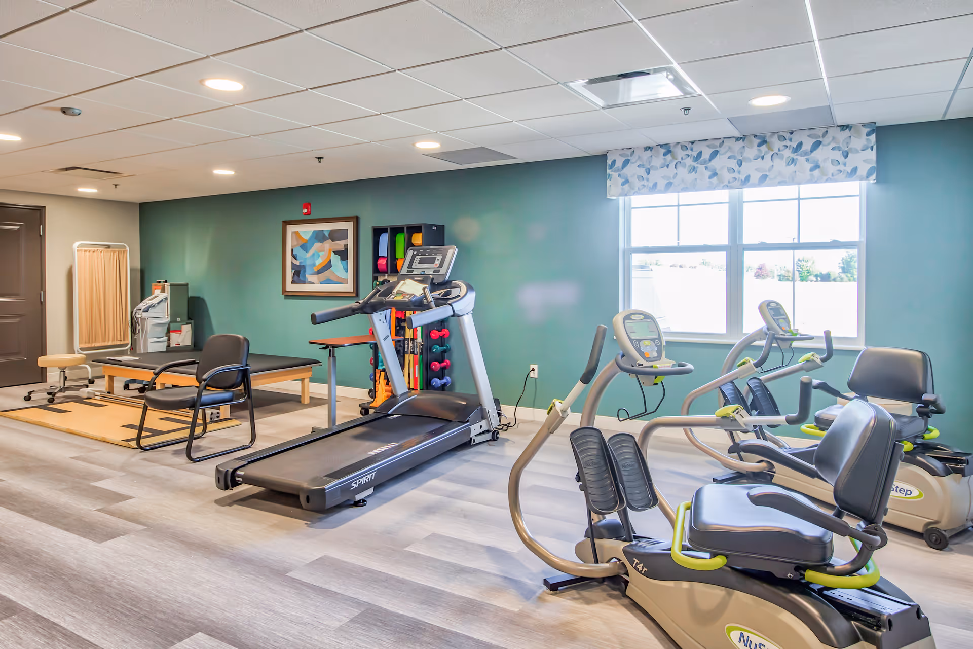 A senior living facility exercise room with a treadmill, two recumbent exercise bikes, a padded treatment table, a chair, and a rack of colorful exercise equipment against a green wall. A window with a patterned valance lets in natural light.