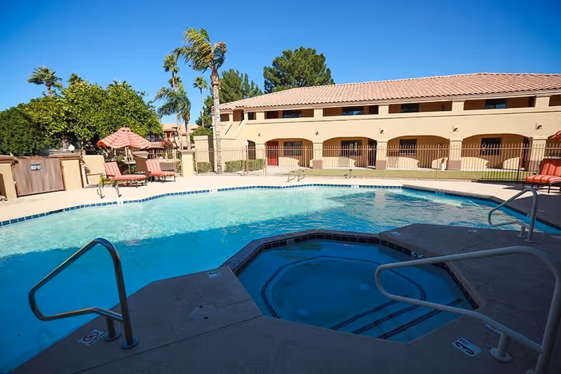 Outdoor swimming pool and hot tub area at a senior living facility with lounge chairs, umbrellas, palm trees, and a beige two-story building in the background under a clear blue sky.