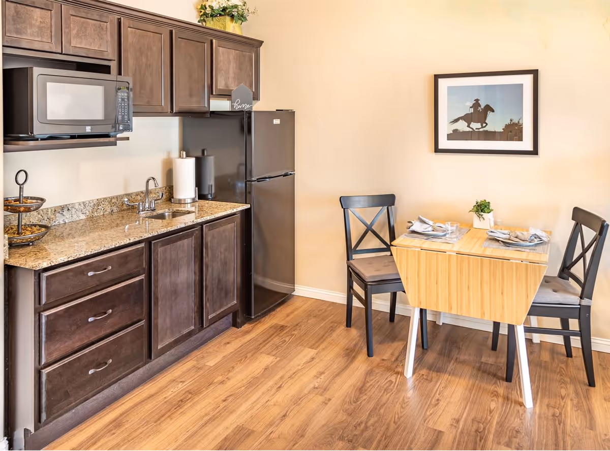 Kitchenette with dark wood cabinets, granite countertop, microwave, refrigerator and a small two-person dining table on wood flooring.