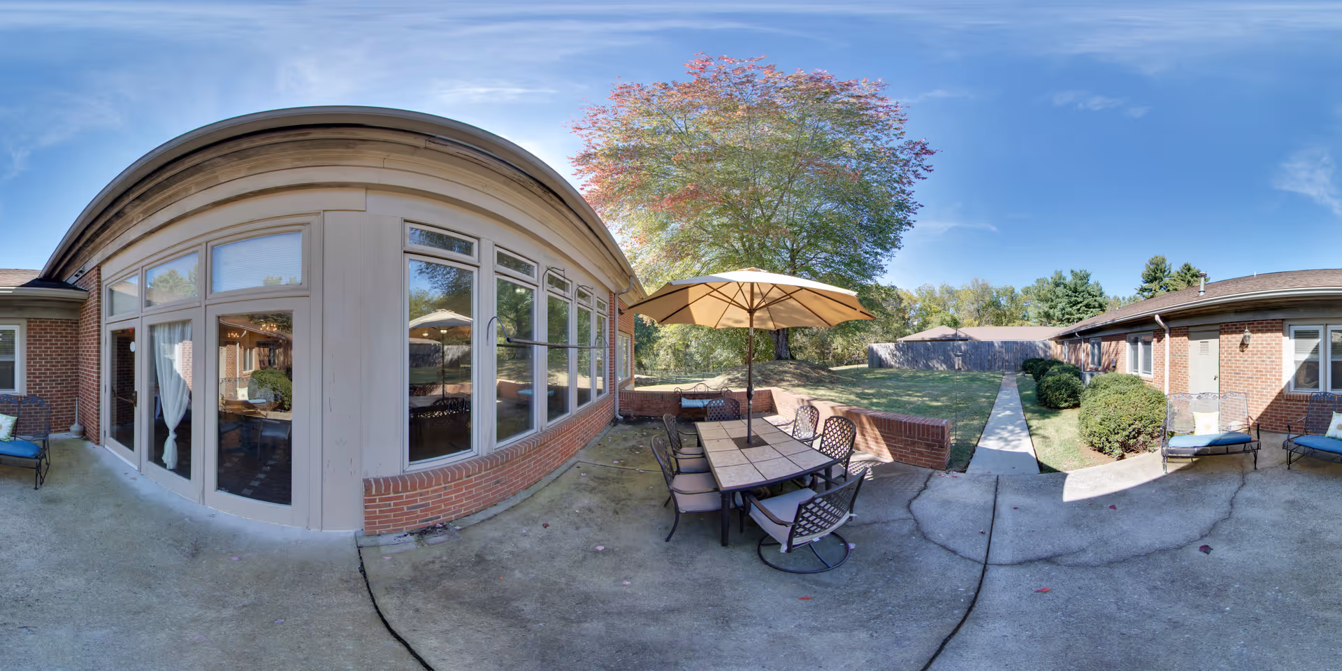 Outdoor patio area at The Elms of Lynchburg featuring a tiled table with six chairs and a large beige umbrella. The patio is surrounded by brick walls and adjacent to a building with large windows and glass doors. There are two lounge chairs on the right side and a concrete walkway leading to a grassy area with trees and a wooden fence in the background under a clear blue sky.