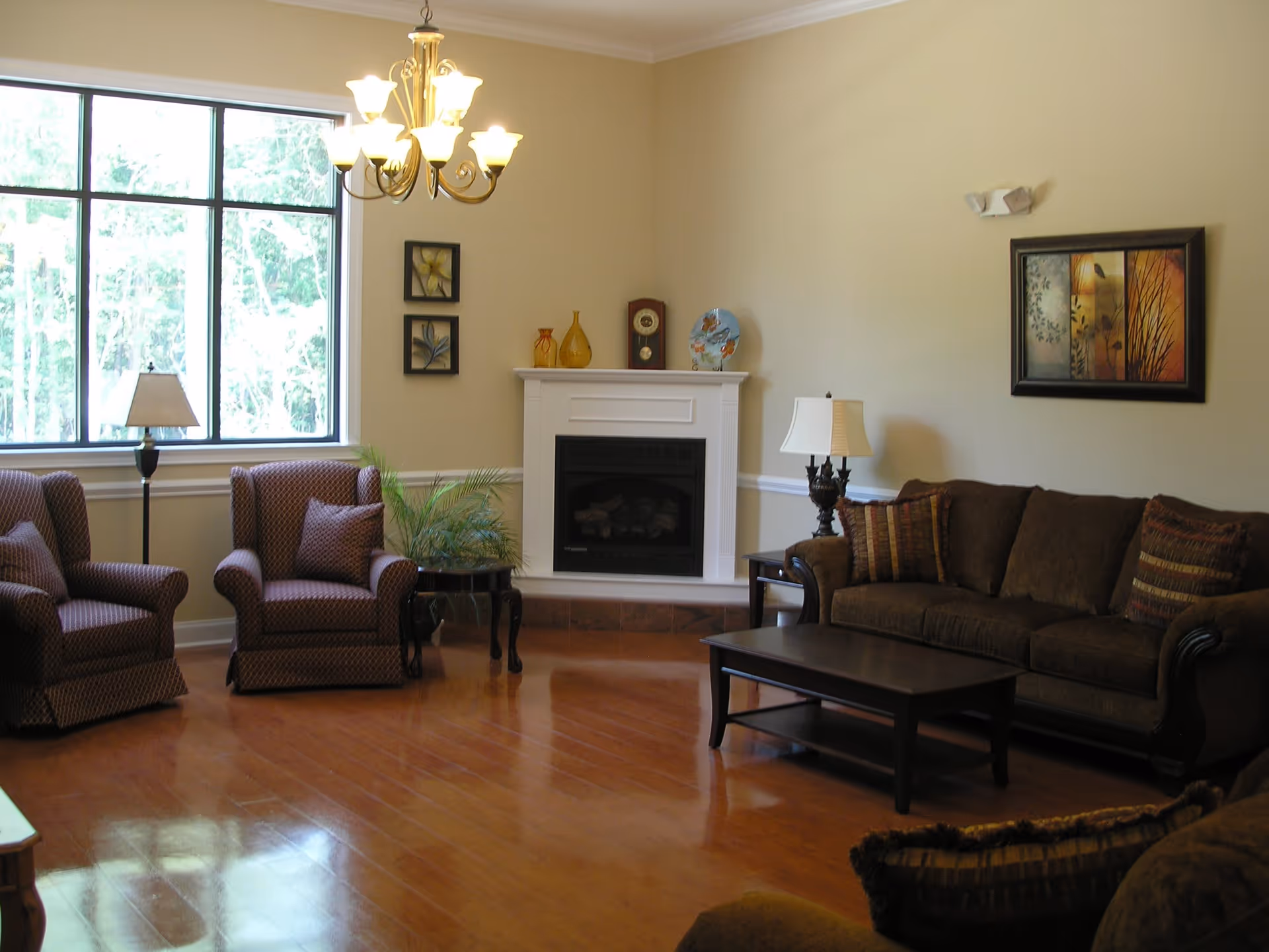 A cozy living room with wooden flooring, a large window letting in natural light, two patterned armchairs, a brown sofa with striped cushions, a dark wooden coffee table, a white fireplace with decorative items on the mantel, two table lamps, and framed artwork on the walls.
