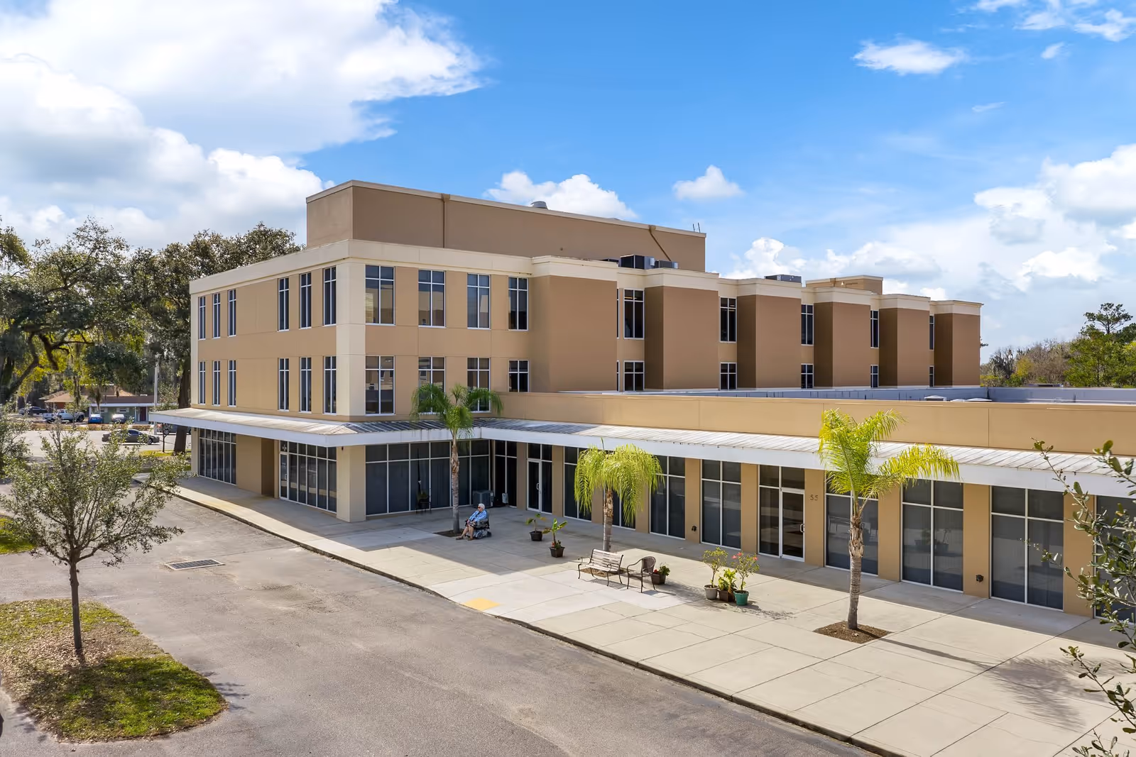 Front exterior of a multi-story beige senior living building with a covered entrance, palm trees, benches and people outside on a sunny day.