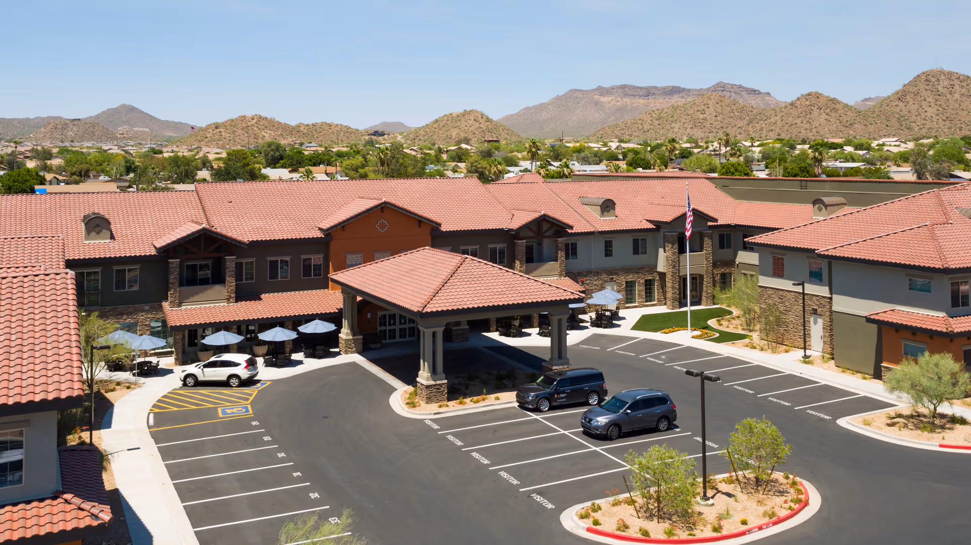 Aerial view of Las Palomas Senior Living facility showing a large building with red-tiled roofs, a covered entrance, parking spaces including handicapped spots, outdoor seating with umbrellas, and surrounding desert landscape with mountains in the background.