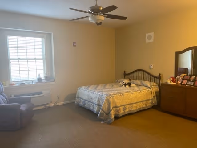 A bedroom with a bed covered in a patterned quilt, a wooden dresser with a mirror and framed photos, a brown armchair near a window with blinds, and a ceiling fan overhead.