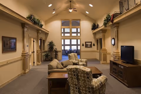 A spacious and warmly lit common area with high vaulted ceilings, featuring a sofa, two patterned armchairs, a wooden coffee table, and a TV on a wooden stand. The room has beige walls with decorative columns and plants placed on ledges near the ceiling. Large windows at the far end let in natural light, and there are framed pictures on the walls.