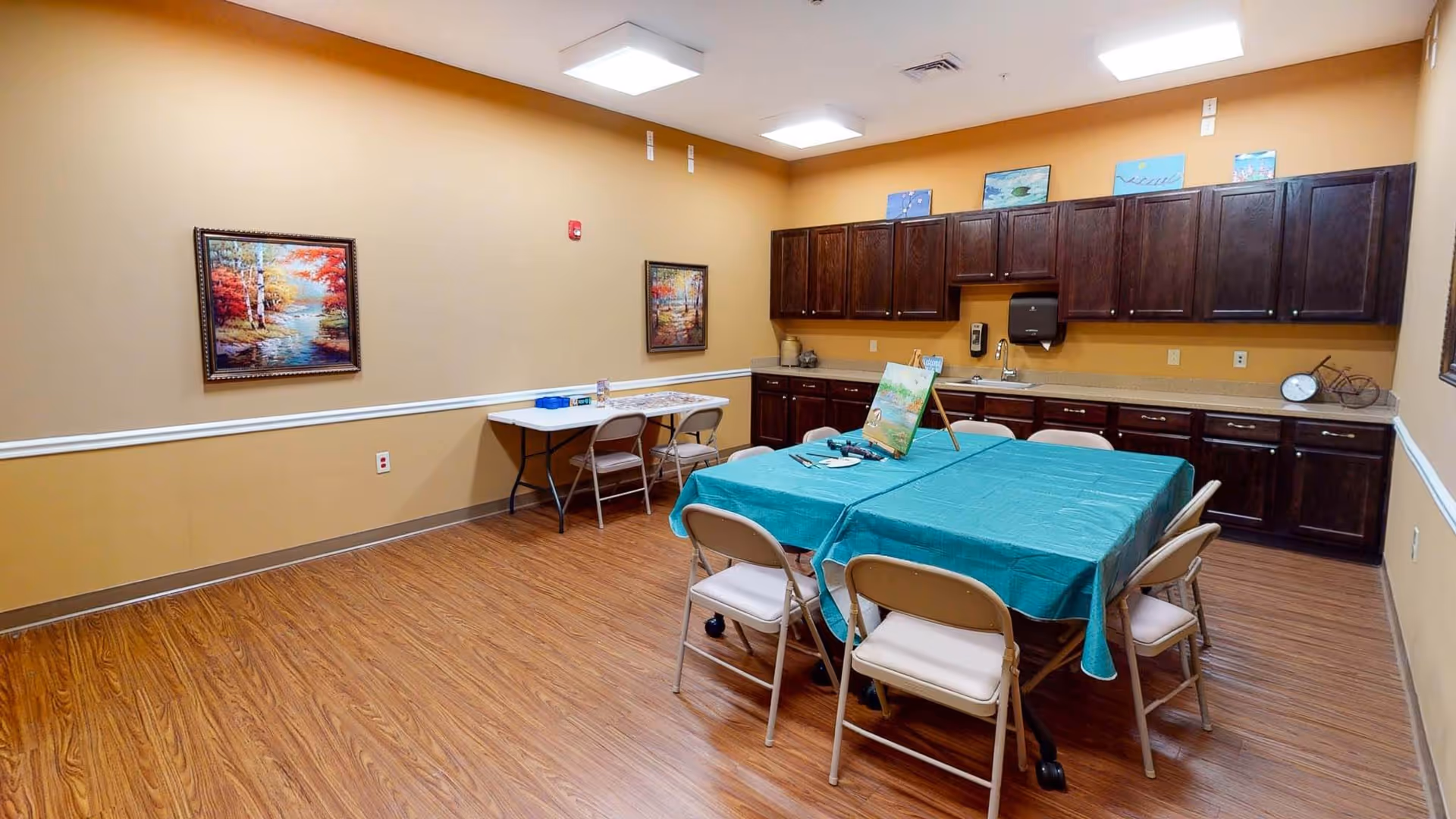 A well-lit activity room with wooden flooring and beige walls. There is a table covered with a teal tablecloth surrounded by folding chairs in the center, with art supplies and a painting on an easel on top. Along the back wall, there are dark wooden cabinets and a countertop with a sink, paper towel dispenser, and decorative items. Two framed landscape paintings hang on the walls, and another folding table with chairs is set against the left wall.