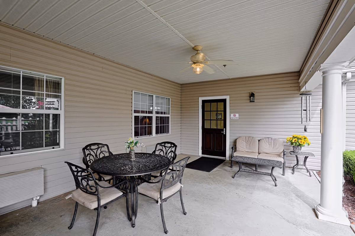 Covered outdoor patio area with a round black metal table and four matching chairs with beige cushions. There is a small vase with flowers on the table. To the right, there is a cushioned bench with a small side table holding a pot of sunflowers. The area has beige siding walls, a ceiling fan with a light, a dark wooden door with glass panels, and two white columns supporting the roof.