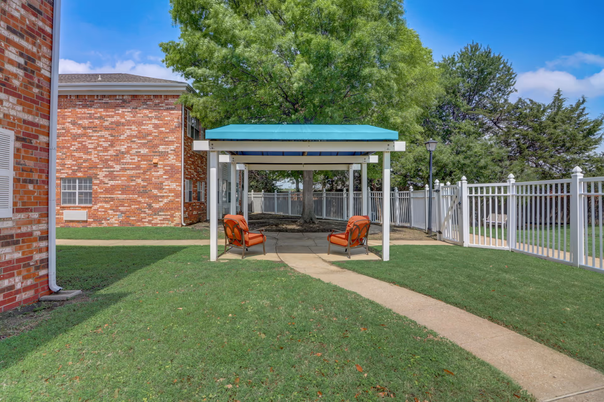 Outdoor seating area with two orange cushioned chairs under a white pergola with a teal roof, situated on a concrete patio surrounded by green grass. A brick building is visible on the left, and a white fence encloses the area with trees in the background under a blue sky with some clouds.