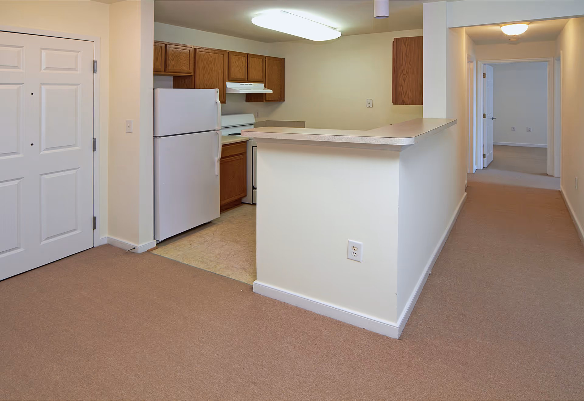 Interior view of an apartment kitchen area with white refrigerator, stove, and wooden cabinets. The kitchen is adjacent to a hallway with beige carpet and white walls, leading to other rooms. A white front door is visible on the left side.