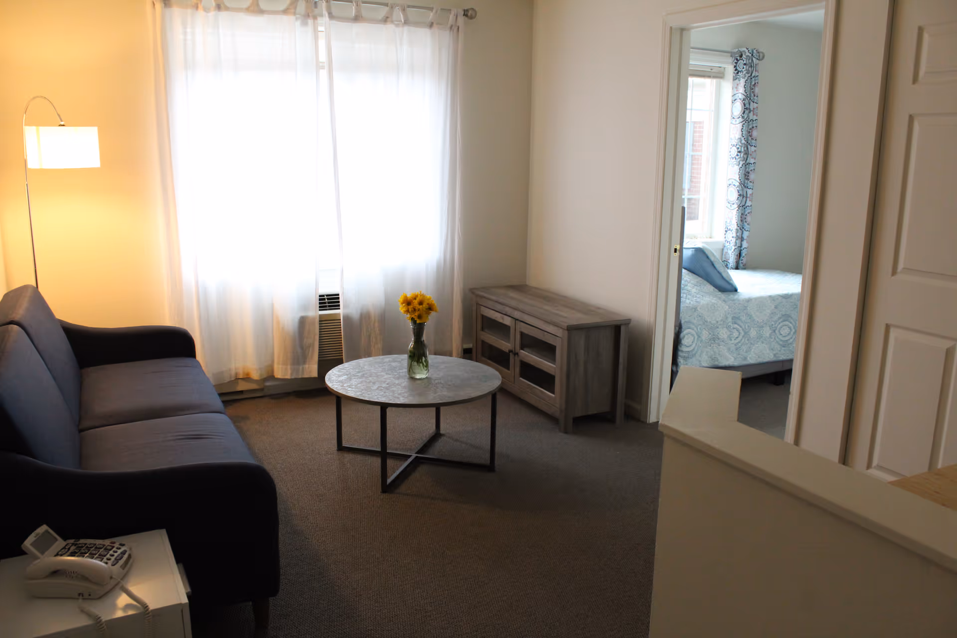 Small furnished living room with a sofa, round coffee table holding a vase of flowers, a TV stand, and a doorway leading to a bedroom.