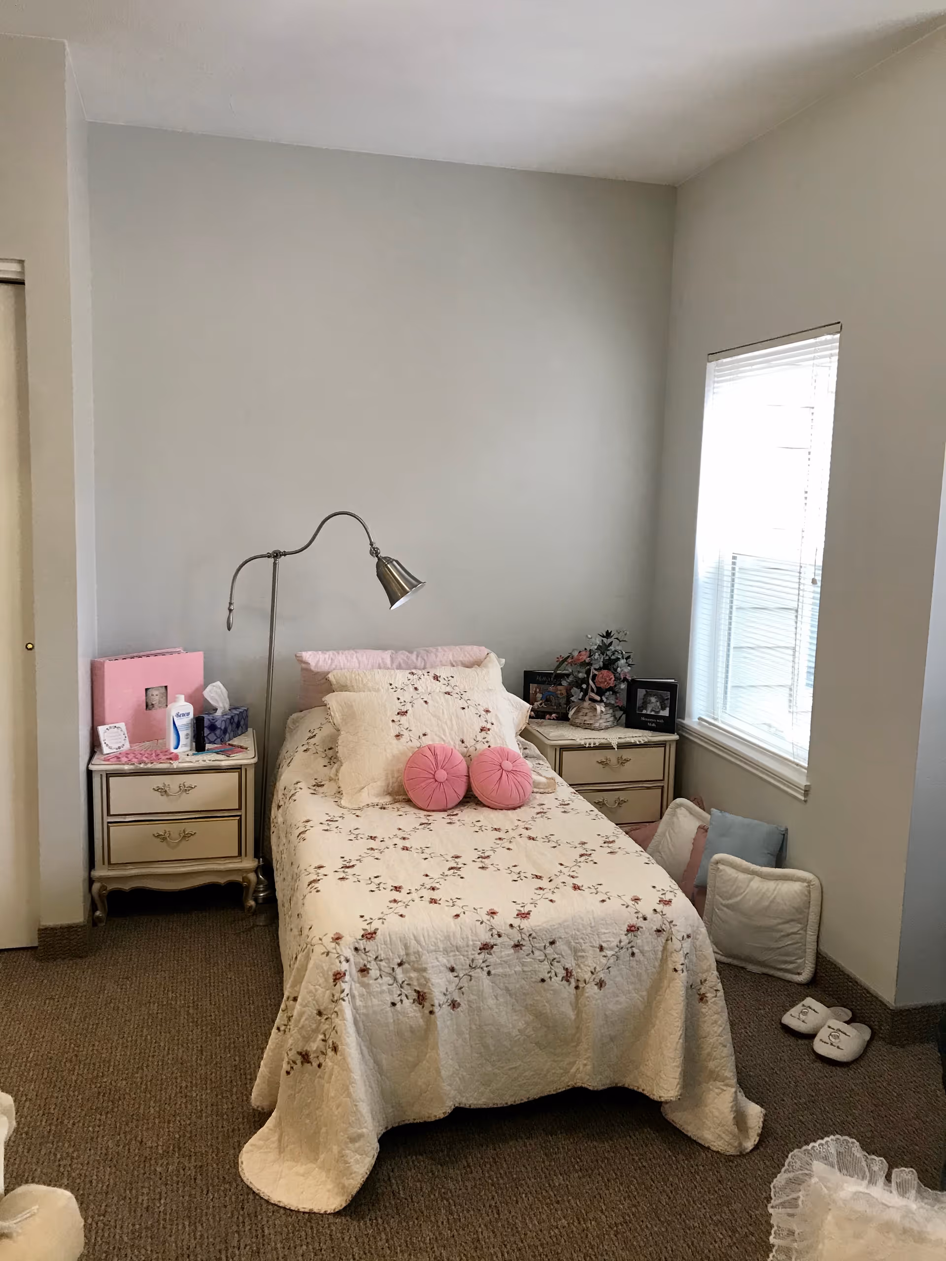 A small bedroom with a floral-covered twin bed, two bedside tables, a floor lamp, and a window.