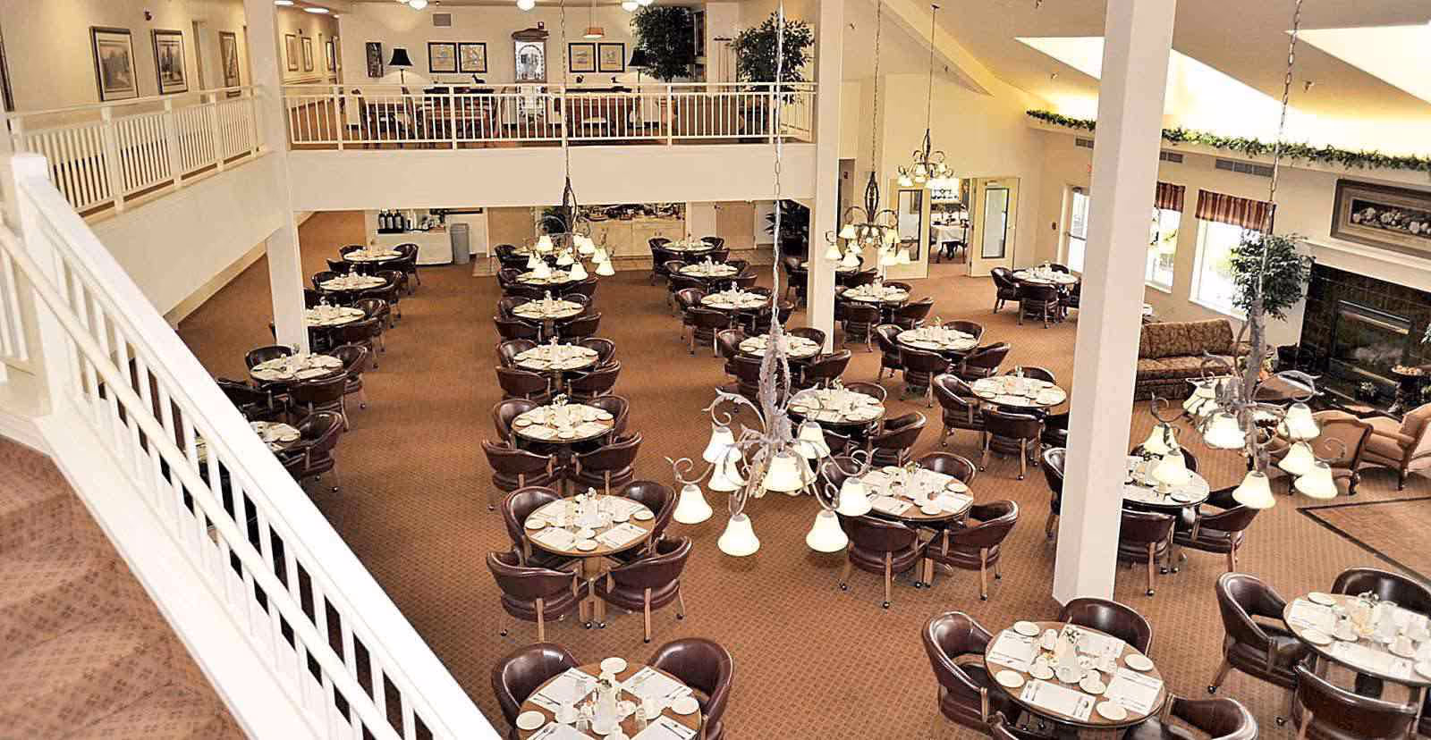 A spacious dining room in a senior living facility with multiple round tables set with plates, cups, and silverware. The room features brown carpet, leather chairs, chandeliers hanging from the ceiling, and a fireplace with seating area on the right side. There is a second-floor balcony overlooking the dining area with additional seating and framed artwork on the walls.