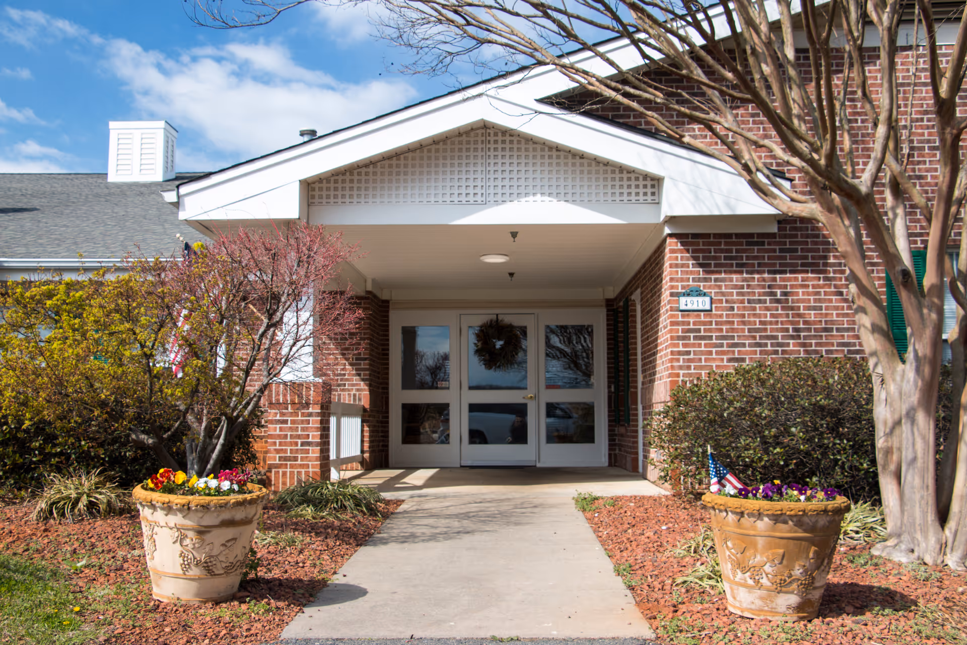Entrance of a brick building with a covered porch, double glass doors decorated with a wreath, two large flower pots with colorful flowers on either side of the walkway, and trees and shrubs surrounding the entrance under a partly cloudy sky.