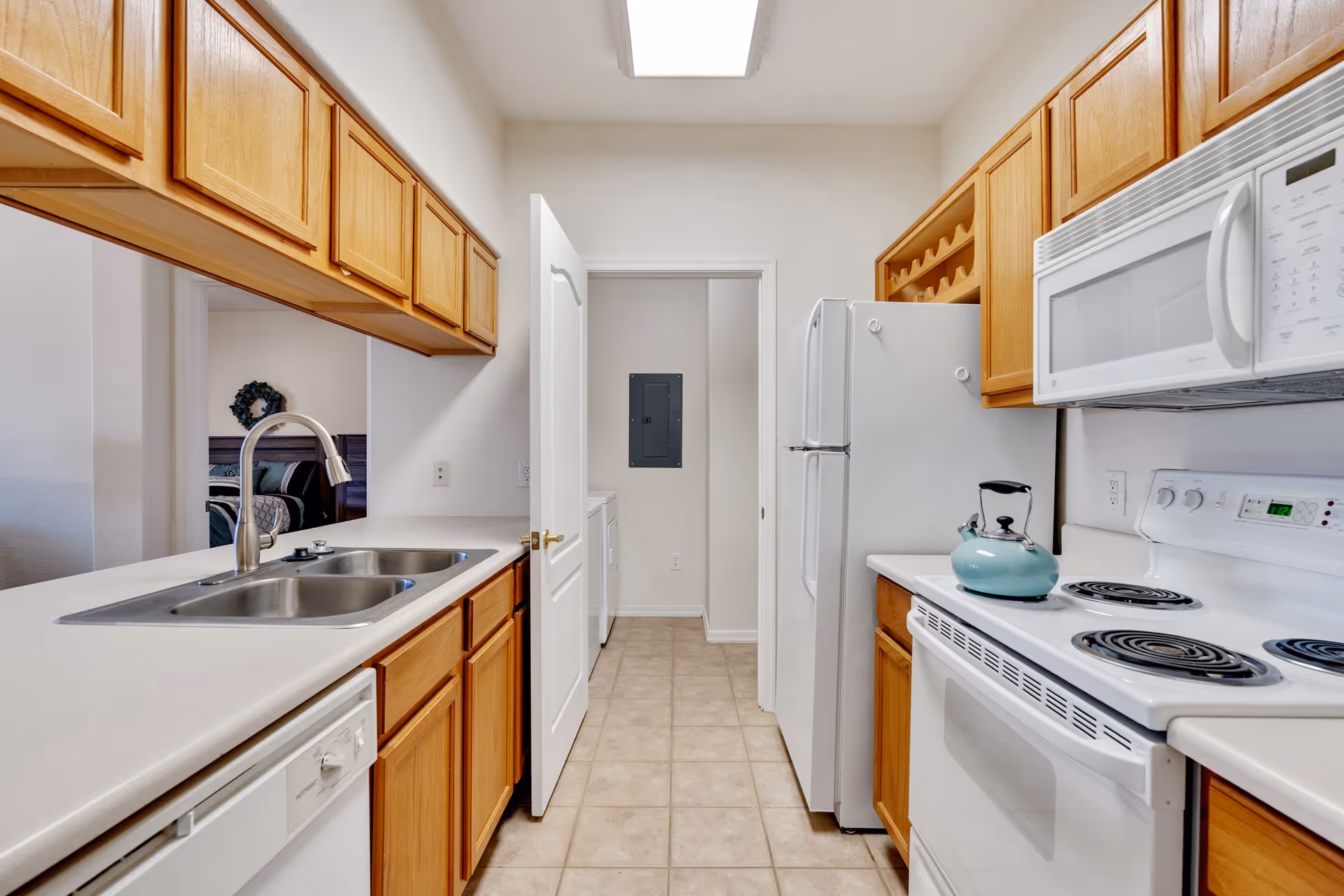A bright kitchen with wooden cabinets, white countertops, and white appliances including a refrigerator, microwave, stove with a teal kettle, and dishwasher. There is a double sink with a modern faucet and a door at the end of the kitchen leading to a laundry area with a washer and dryer. A glimpse of a bedroom is visible through an open space on the left.