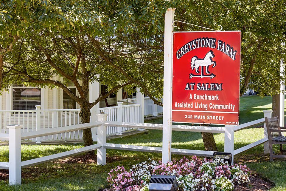 Outdoor view of Greystone Farm at Salem assisted living community sign with a white fence, green grass, blooming flowers, trees, and part of the building with a porch in the background.