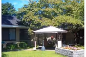 Single-story health care building with a small canopy gazebo over a courtyard seating area, surrounded by lawn, trees, and a stone planter.