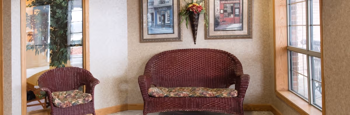A cozy seating area with a burgundy wicker loveseat and matching chair, both with floral cushions. The wall behind features two framed pictures and a decorative wall-mounted flower arrangement. To the right, there is a large window with wooden trim letting in natural light, and to the left, a glass door with a view of a plant and outdoor area.