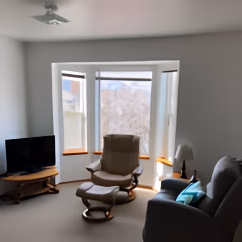 A bright living room with a recliner and ottoman in front of a bay window, a sofa with pillows, and a TV on a wooden stand.