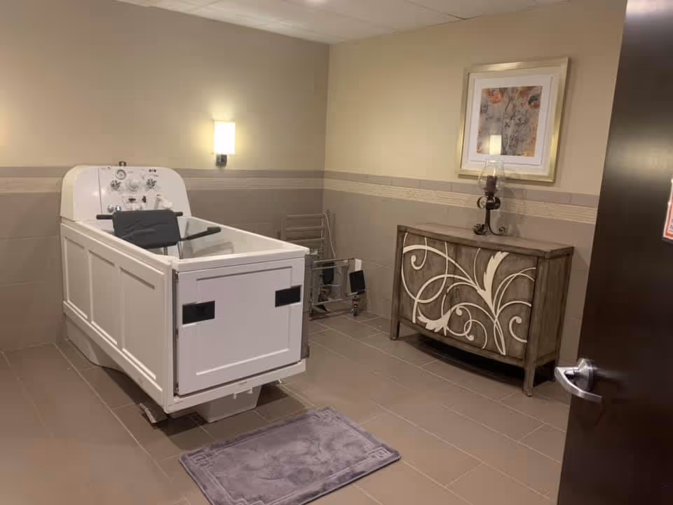 A room with a large white assisted bathing tub equipped with controls and a seat, a small decorative cabinet with a candle holder and framed artwork above it, a wall-mounted light fixture, and a purple bath mat on the tiled floor.
