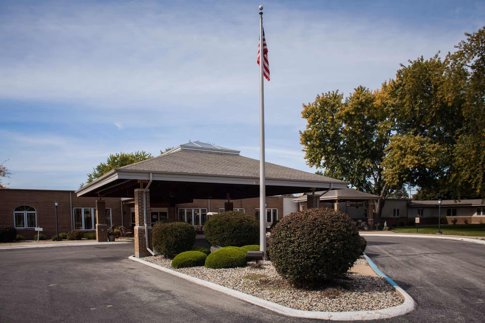 Front entrance of Living Heritage Village with a covered porte-cochere, central flagpole, trimmed bushes, and a brick building under a blue sky.