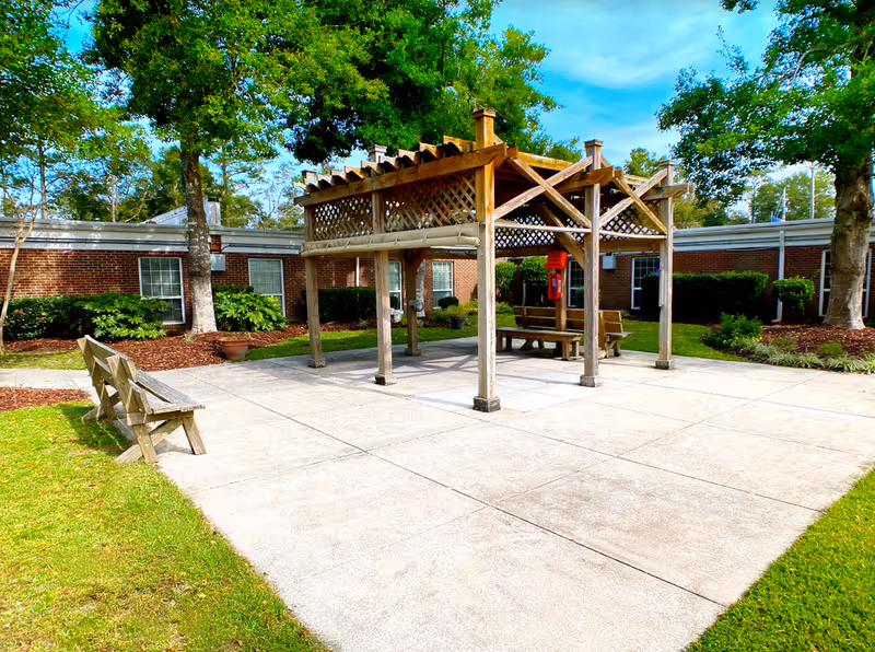 Outdoor courtyard area at Cypress Pointe Rehabilitation Center featuring a wooden pergola with benches underneath, surrounded by green grass, trees, and shrubs, with a brick building in the background under a partly cloudy blue sky.